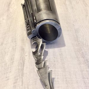 May include: A metallic gray prop weapon with a cylindrical body and a jagged blade. The cylindrical body has a rectangular window and a ribbed section. The blade is composed of several stylized, pointed elements.