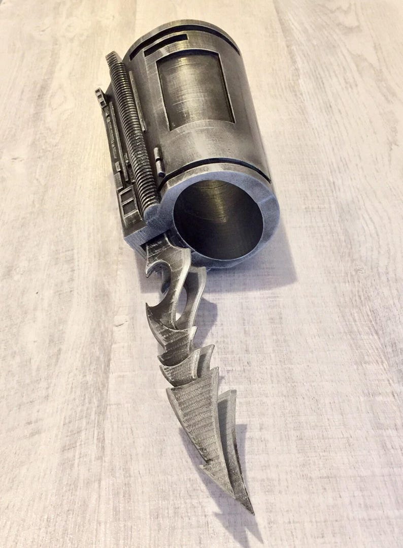 May include: A metallic gray prop weapon with a cylindrical body and a jagged blade. The cylindrical body has a rectangular window and a ribbed section. The blade is composed of several stylized, pointed elements.