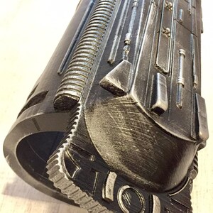 May include: A detailed, metallic-looking cylindrical object with intricate, raised patterns and textures. The color scheme is primarily dark gray with silver highlights, giving it a weathered appearance. The design includes a series of parallel lines, geometric shapes, and what appears to be a gear-like edge.