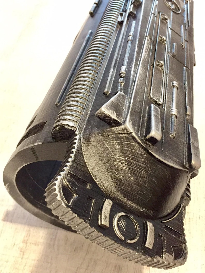 May include: A detailed, metallic-looking cylindrical object with intricate, raised patterns and textures. The color scheme is primarily dark gray with silver highlights, giving it a weathered appearance. The design includes a series of parallel lines, geometric shapes, and what appears to be a gear-like edge.