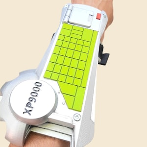 May include: Silver and green wrist-mounted device with the text "XP9000" on a circular component. The device features a grid of green squares and a red button. The design is futuristic and mechanical.