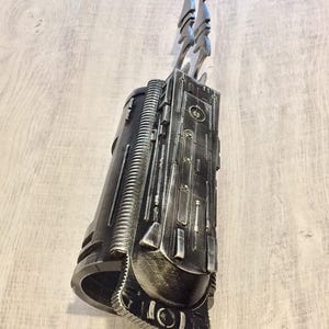 May include: A metallic gray prop weapon with a cylindrical body and intricate detailing. Two stylized blades extend from the top. The prop has a weathered appearance, suggesting a futuristic or sci-fi theme. The design incorporates various geometric shapes and textures.