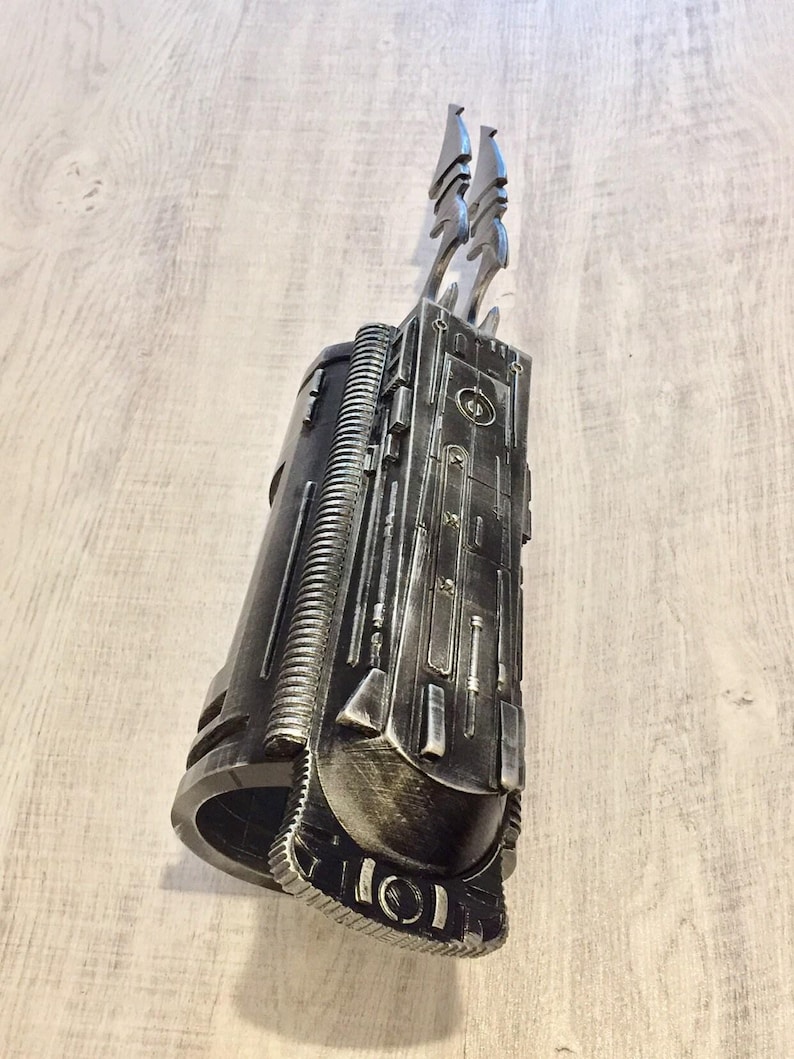 May include: A metallic gray prop weapon with a cylindrical body and intricate detailing. Two stylized blades extend from the top. The prop has a weathered appearance, suggesting a futuristic or sci-fi theme. The design incorporates various geometric shapes and textures.