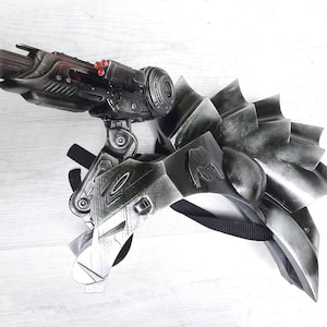 Pode incluir: Uma arma de adereço futurista, prateada e preta, com uma lente azul e luzes vermelhas. O adereço tem um design complexo com múltiplos componentes e uma correia preta. A estética geral sugere um tema de ficção científica ou cosplay.