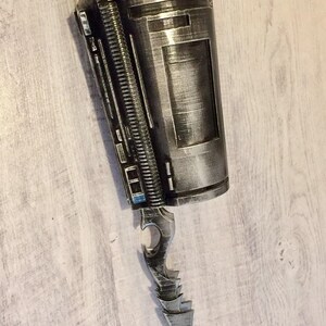 May include: A silver and black prop weapon with a cylindrical body and a blade-like extension. The design features intricate details, including a ridged spine and a sharp, pointed blade. The prop has a metallic finish and appears to be a futuristic or fantasy-themed item.