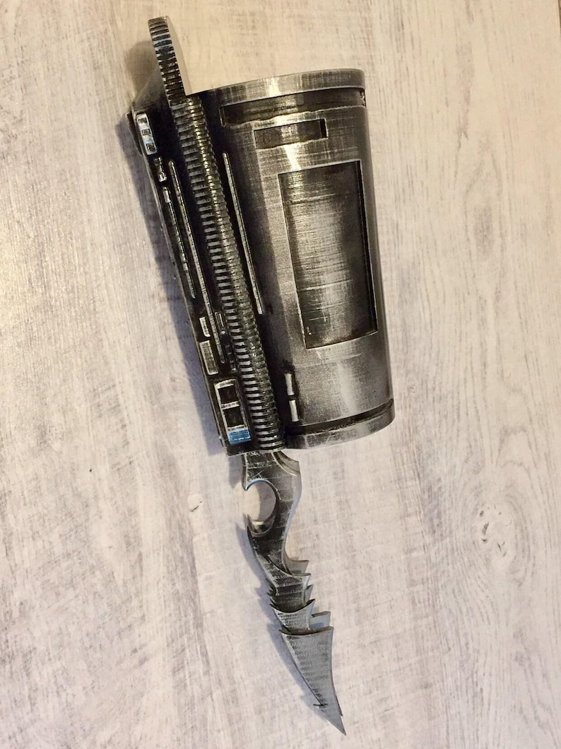May include: A silver and black prop weapon with a cylindrical body and a blade-like extension. The design features intricate details, including a ridged spine and a sharp, pointed blade. The prop has a metallic finish and appears to be a futuristic or fantasy-themed item.