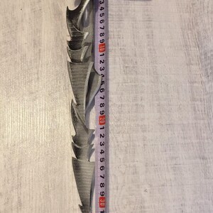May include: A gray, jagged-edged blade, approximately 12 inches long, with a decorative handle. A measuring tape is placed next to the blade, showing its length. The blade has a series of pointed, curved edges.