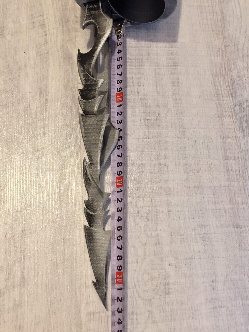 May include: A gray, jagged-edged blade, approximately 12 inches long, with a decorative handle. A measuring tape is placed next to the blade, showing its length. The blade has a series of pointed, curved edges.