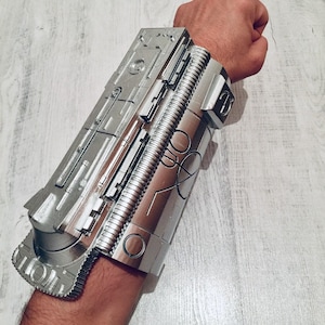 May include: Silver-colored, futuristic-looking wrist armor with intricate mechanical details. The armor is made of a metallic material and features various panels, ridges, and circular designs. The design suggests a science fiction or cosplay theme.