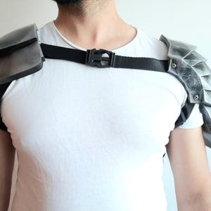 May include: Silver-coloured shoulder armour pieces, connected by a black strap with a buckle. The armour has a weathered appearance, with layered plates on one side and a smooth, curved design on the other. The armour is worn over a white t-shirt.