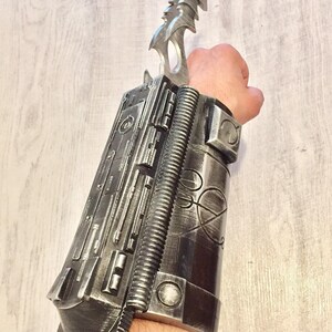 May include: A silver-colored wrist-mounted prop with a blade. The prop features a detailed, mechanical design with various textures and shapes, including a ribbed section and a pointed blade extending upwards. The overall aesthetic suggests a futuristic or science fiction theme.
