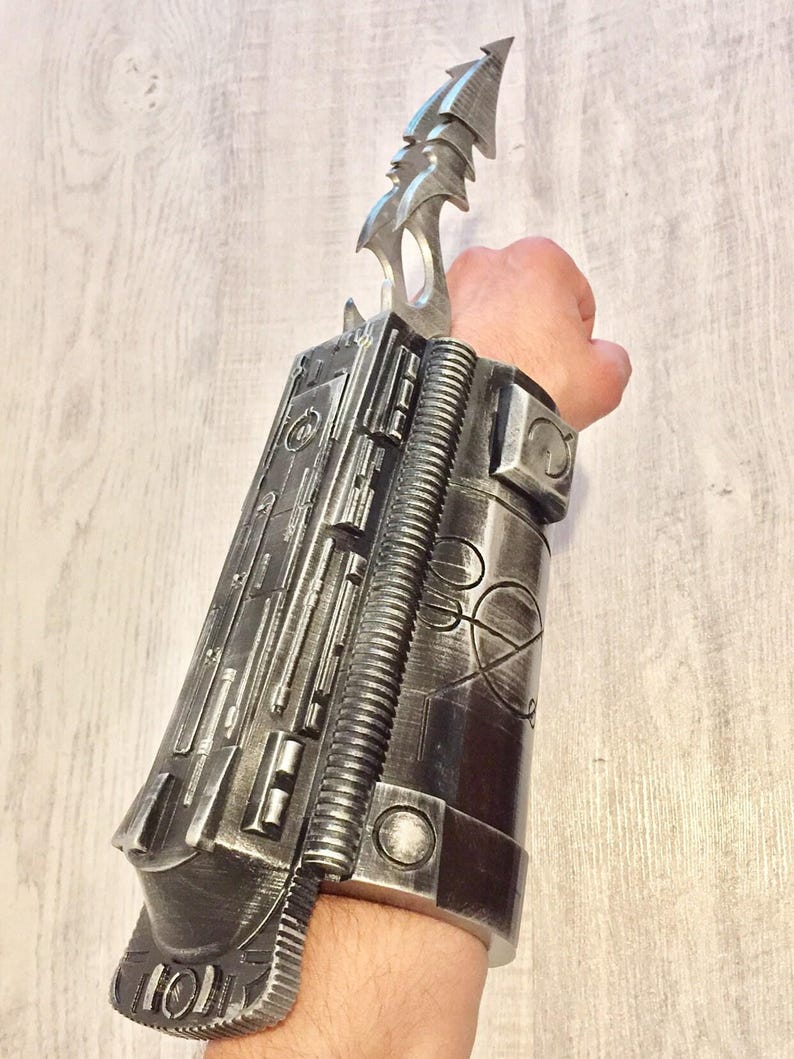 May include: A silver-colored wrist-mounted prop with a blade. The prop features a detailed, mechanical design with various textures and shapes, including a ribbed section and a pointed blade extending upwards. The overall aesthetic suggests a futuristic or science fiction theme.