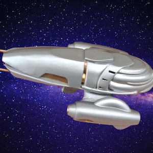Puede incluir: Maqueta de nave espacial plateada con detalles dorados, sobre un fondo de galaxia estrellada. La nave espacial presenta un cuerpo redondeado, paneles detallados y elementos extendidos. Un artículo futurista con temática espacial.