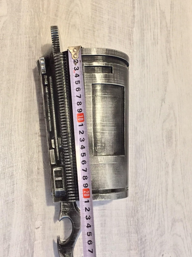May include: A silver-colored, cylindrical prop with a rectangular cutout and intricate detailing. A measuring tape shows the prop is approximately 9 inches in length. The prop has a hook-like extension at the bottom.