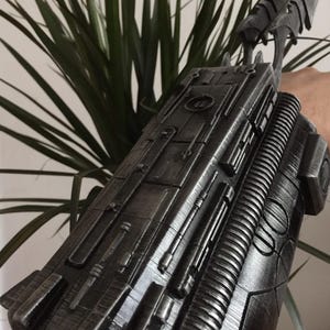 May include: A metallic gray arm gauntlet with intricate detailing, featuring raised panels, cylindrical elements, and a ribbed section. The gauntlet has a futuristic design with a series of blade-like protrusions extending from the top.