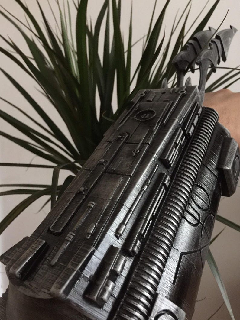 May include: A metallic gray arm gauntlet with intricate detailing, featuring raised panels, cylindrical elements, and a ribbed section. The gauntlet has a futuristic design with a series of blade-like protrusions extending from the top.