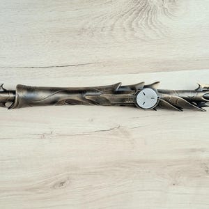 Peut inclure: Une arme fantastique en bronze et argent, peut-être un sabre laser ou un accessoire similaire. Le corps central présente un design texturé et nervuré avec un élément circulaire. Les extrémités présentent des détails complexes en forme de griffes.