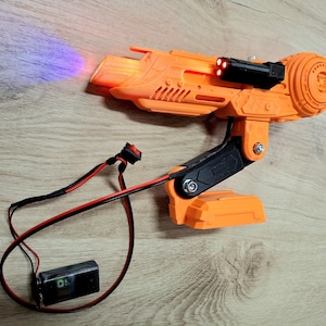 Pode incluir: Uma arma de raios futurista laranja e preta com um feixe de luz roxa que emana do cano. A arma tem uma alça preta e um pacote de bateria conectado por fios vermelhos e pretos. A arma tem três luzes vermelhas na parte superior.
