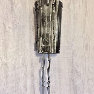 May include: A detailed, metallic sculpture with a cylindrical body and intricate surface detailing. Two curved, pointed blades extend from the bottom. The piece has a futuristic, industrial aesthetic.