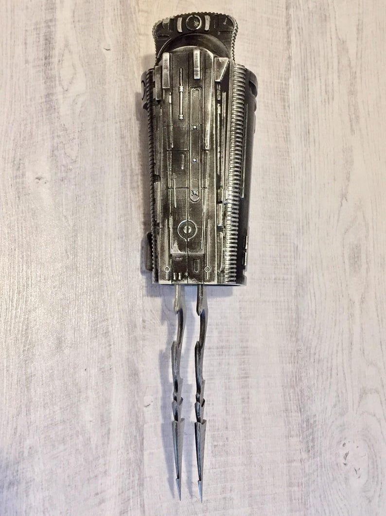 May include: A detailed, metallic sculpture with a cylindrical body and intricate surface detailing. Two curved, pointed blades extend from the bottom. The piece has a futuristic, industrial aesthetic.