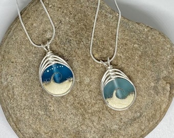 May include: Two silver chain necklaces with teardrop-shaped pendants. Each pendant features a blue and white resin design with a wave and sand.