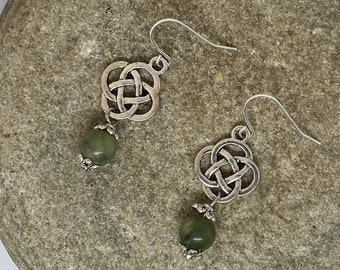 May include: Silver Celtic knot earrings with green stone beads. The earrings are hanging from a branch on a rock.