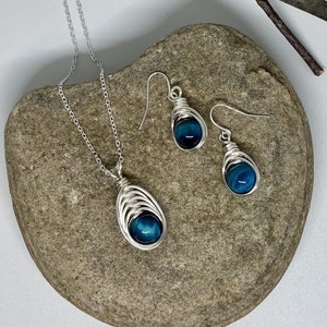 May include: A silver chain necklace and matching earrings with blue tiger's eye stones wrapped in silver wire. The jewellery is displayed on a grey rock.