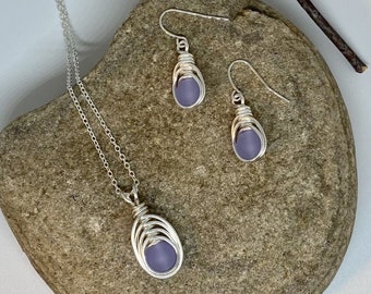 May include: A silver necklace and earrings set with a lavender glass pendant and matching earrings. The jewelry is displayed on a gray rock with a branch in the background.
