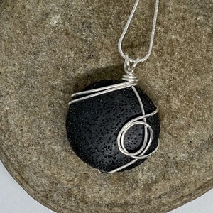 May include: A black lava rock pendant wrapped in silver wire, hanging from a silver chain. The pendant is on a gray rock background.
