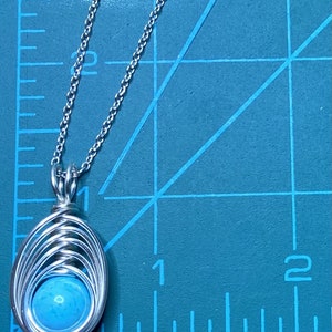 Op de afbeelding: Een zilveren ketting met een hanger van turquoise steen. De hanger is gemaakt van draad dat in een cirkelvorm om de steen is gewikkeld.