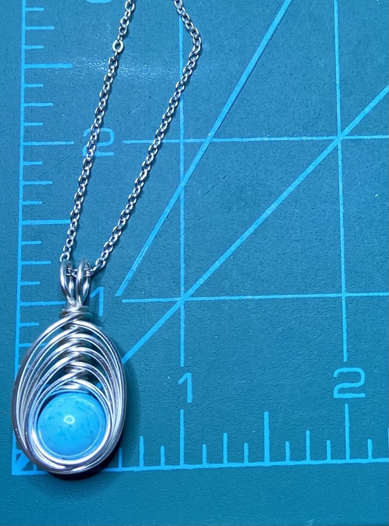 Op de afbeelding: Een zilveren ketting met een hanger van turquoise steen. De hanger is gemaakt van draad dat in een cirkelvorm om de steen is gewikkeld.