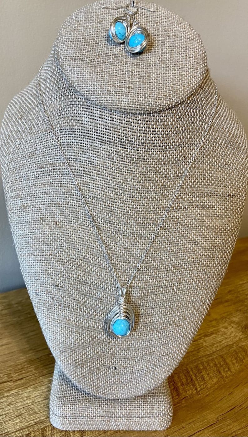 Op de afbeelding: Een turquoise stenen ketting en oorbellen set met zilveren draadwikkeling. De ketting heeft een hanger met een turquoise steen gewikkeld in zilveren draad. De oorbellen zijn ook gemaakt met turquoise stenen gewikkeld in zilveren draad.