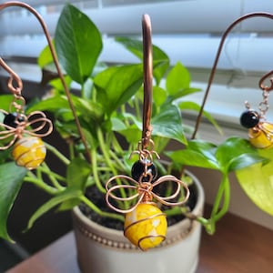 May include: Copper wire earrings with a bee design. Each earring features a yellow and white striped bead, a black bead, and copper wire wings. The earrings are suspended from a copper wire hook.