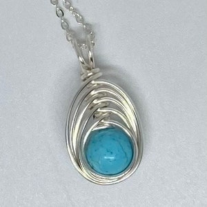 Op de afbeelding: Een zilveren ketting met een hanger van turquoise steen. De hanger is gewikkeld in zilverdraad.