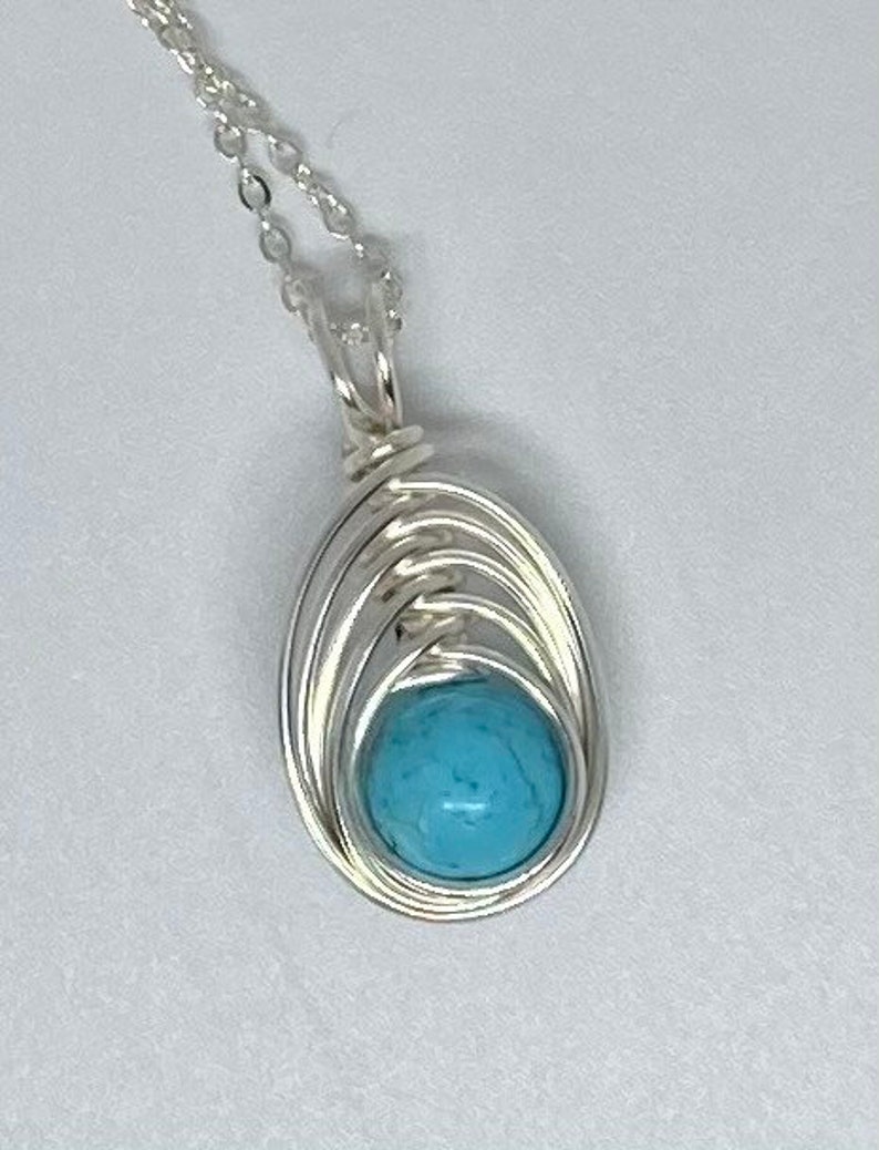 Op de afbeelding: Een zilveren ketting met een hanger van turquoise steen. De hanger is gewikkeld in zilverdraad.