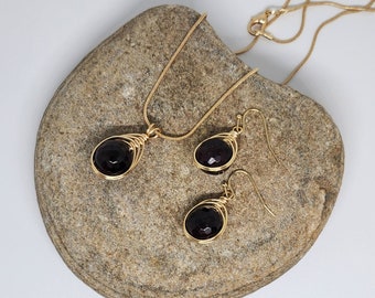 May include: A gold necklace and earrings set with a dark red gemstone pendant and matching earrings. The jewelry is displayed on a light brown rock.