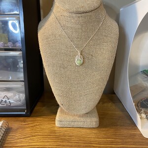 May include: A silver chain necklace with a green stone pendant on a burlap jewelry display stand.