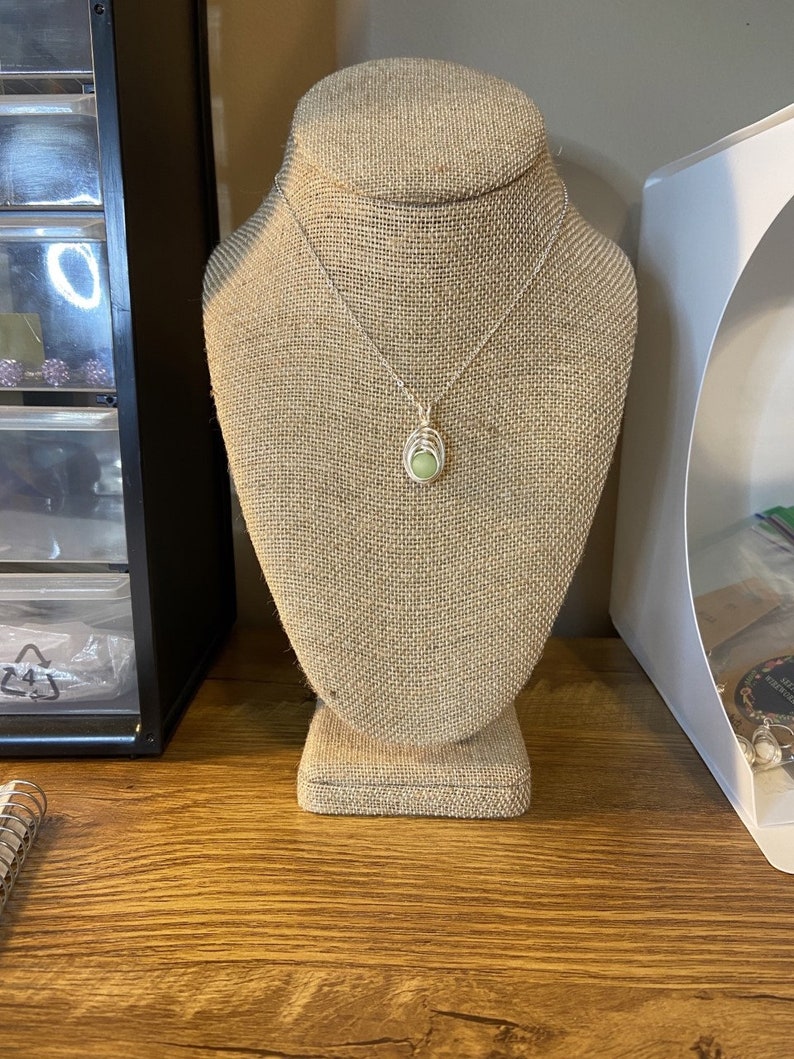 May include: A silver chain necklace with a green stone pendant on a burlap jewelry display stand.