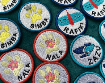 May include: A collection of colorful embroidered iron-on patches featuring animal paw prints and names like Simba, Nala, and Rafiki. The patches are circular and have a white background with colorful embroidery.