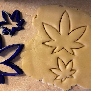 May include: A close-up shot shows blue cookie cutters in the shape of cannabis leaves next to a piece of dough with leaf imprints. The dough is light brown, and the cutters are a deep blue.