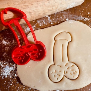 Puede incluir: Un cortador de galletas de plástico rojo con forma de pene junto a un trozo de masa con la forma de pene recortada. Un rodillo de madera está en el fondo.