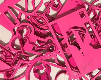May include: A collection of bright pink cookie cutters in various shapes, including a guitar, a key, and a person sitting. The cutters are made of plastic and are scattered across a surface.