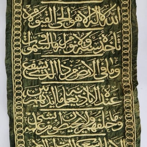 Op de afbeelding: Een groene stoffen banner met gouden borduurwerk van Arabische tekst. De banner heeft een decoratieve rand van gouden cirkels. De tekst bevat de zin "Bismillah ir-Rahman ir-Rahim" en "La ilaha illa-llah Muhammadur Rasulullah".