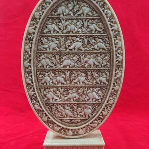 May include: A white oval-shaped decorative plaque with intricate carvings of elephants in a procession. The plaque is mounted on a white pedestal with similar carvings.