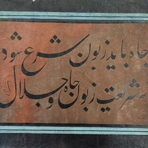 May include: A framed piece of calligraphy in black ink on a brown background. The calligraphy is in Arabic script and reads "Jaana Baazaroon"