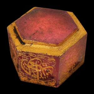 Puede incluir: Una caja hexagonal de estilo antiguo con un cuerpo rojo translúcido y adornos dorados. La tapa también es hexagonal. Los lados de la caja presentan diseños dorados ornamentados. La caja está sobre un fondo negro.