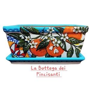 Può includere: Un vaso rettangolare in ceramica con un bordo blu e uno sfondo bianco. Il vaso è decorato con un disegno dipinto a mano di fiori d'arancio e arance. Il vaso ha un sottovaso abbinato. Il testo "La Bottega dei Pincisanti" è stampato sotto il vaso.