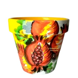 May include: A yellow terracotta flower pot with a hand-painted design of red pomegranates and green leaves. The pot has a white background and the text "La Bottega dei Pincisanti" is written in black on the side.