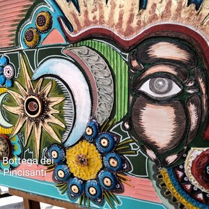 Può includere: Un'opera d'arte colorata in tecniche miste che presenta un grande occhio, un sole, lune e fiori. L'opera d'arte è intitolata "La Bottega del Pincisanti".