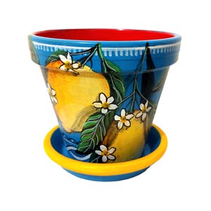 Peut inclure: Pot de fleur en céramique peint à la main avec une soucoupe assortie. Le pot est peint avec un fond bleu et présente des citrons jaunes, des feuilles vertes et des fleurs blanches. Le bord est rouge et la soucoupe a un bord jaune.